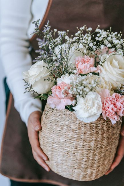 A person holding a woven wicker basket filled with an arrangement of fresh flowers, including white roses, pink carnations, and delicate baby's breath, accented with sprigs of lavender and greenery. T